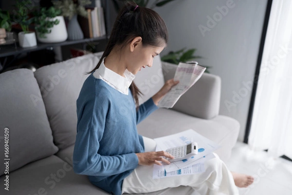 Fototapeta Asian woman holding paper document calculating money savings paying bills in mobile application using smart phone payment online with paying household bills tax insurance managing budget