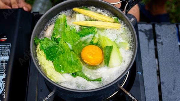Fototapeta Close-up of boiling pot filled with fresh leafy greens baby corn and raw egg cooking in clear broth over portable gas stove.