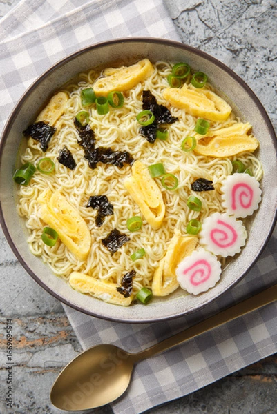 Obraz Saimin is a Hawaiian noodle soup consisting of soft wheat egg noodles served in a hot dashi garnished with diced green onions, kamaboko and omelette closeup in the plate. Vertical top view from above
