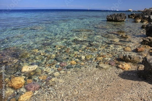 Fototapeta Clear, shallow water laps a pebbled beach, bordered by dark rocks, under a bright sky