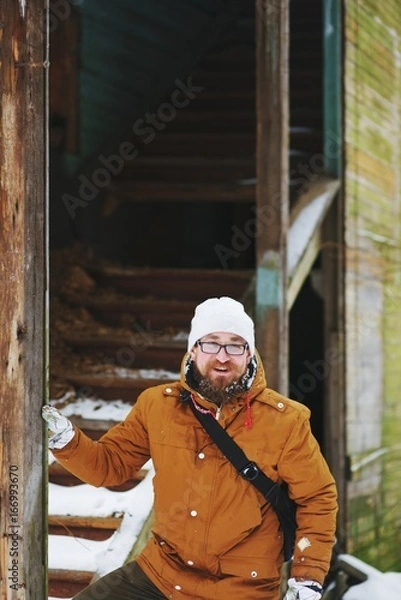 Fototapeta Young smiling red-bearded man stands near the old abandoned houses while traveling in the winter.