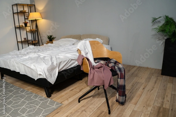 Fototapeta In a stylish bedroom filled with soft light, a bed stands unmade while clothes hang from a chair, reflecting a comfortable, carefree start to the day.