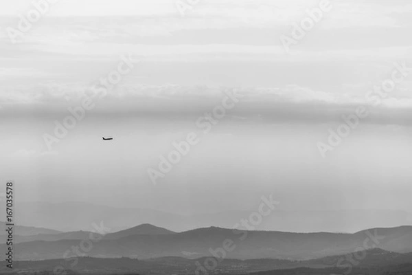Fototapeta A distant airplane flying over some mountains and hills in the middle of mist