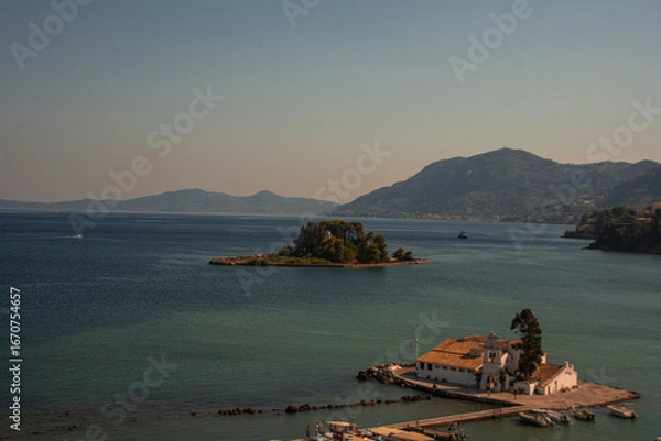 Fototapeta Vlacherna Monastery, located on a small islet near Corfu, Greece, is seen in the foreground with its iconic white buildings and red-tiled roof. The calm blue waters of the Ionian Sea surround it. In t