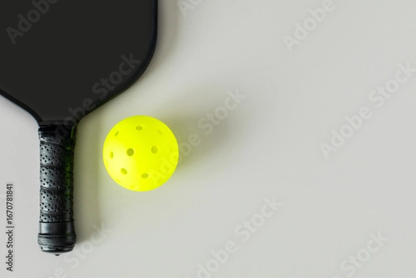Fototapeta Close-up of a black pickleball paddle and bright yellow ball on light neutral background, photographed from above with copy space..
