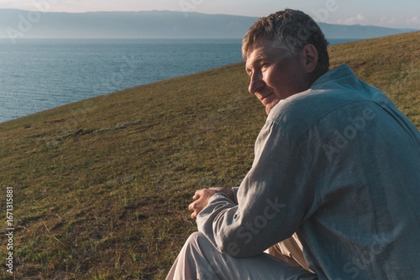 Fototapeta candid portrait of a man enjoys moment tranquility while seated on grassy slope. he gazes thoughtfully across serene lake, basking in warm light setting sun. close up.