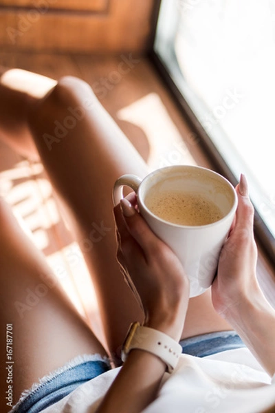 Fototapeta Young woman in smart watch sits by the window with a cup of coffee in her hands on a sunny day