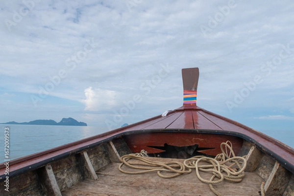 Fototapeta View from the bow of a traditional longtail boat on calm water, with distant islands under a cloudy sky, evoking tropical travel and adventure.