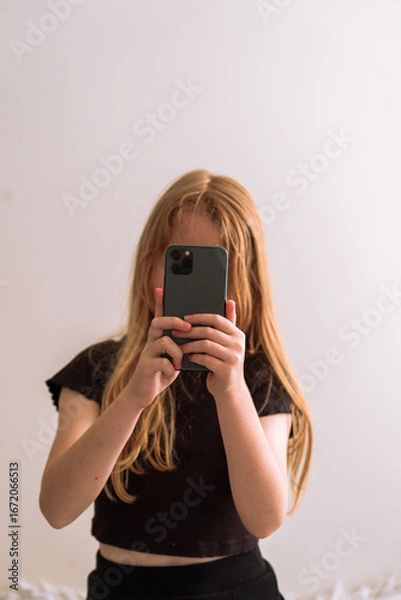 Fototapeta Young girl taking a selfie with a smartphone, standing in front of a light wall. Minimalist and candid lifestyle moment with soft natural lighting.