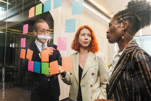 Fototapeta Diverse team collaborates in modern office, brainstorming with sticky notes on glass wall. Startup workshop highlights strategy, planning, leadership, teamwork, and creative meeting energy.