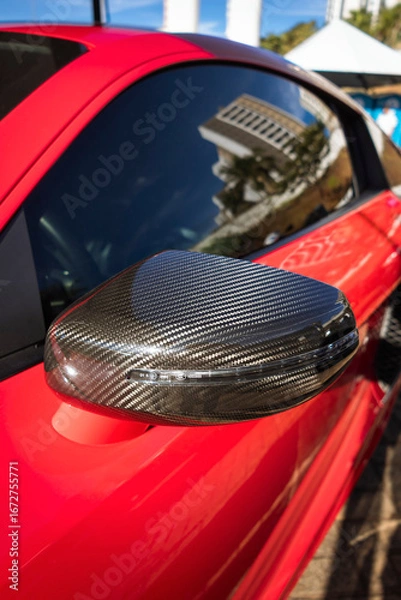 Fototapeta Car side mirror with a carbon fiber cover and integrated turn signal repeater, on a vehicle with red paint.