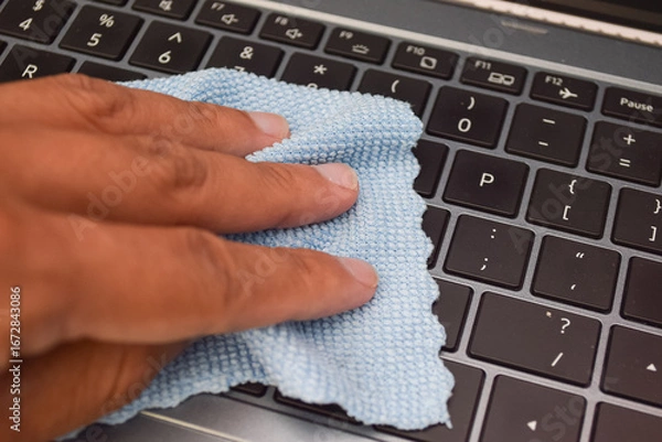 Fototapeta Hand cleaning laptop keyboard with blue microfiber cloth, concept of hygiene, device care, and electronic maintenance.