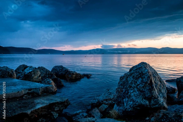 Fototapeta Large blocks of stones on the shore of the lake against the backdrop of a beautiful sunset