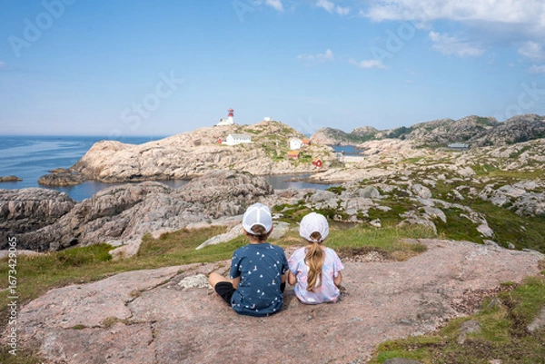 Obraz Two small children sitting on a rock looking at the Lindesnes Lighthouse. Lindesnes Fyr on most southern point of Norway, Europe. 