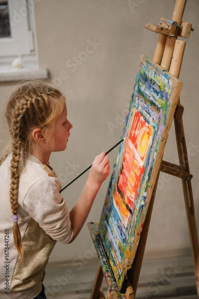 Fototapeta Child draw a picture in art class. Girl enjoys painting in a workshop. Art lesson. Education and learning concept. School kid drawing with brush landscapes on easel. Mix and combine colors.