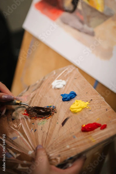 Fototapeta Woman painting in a workshop. Girl mixing and combining colors. Female is drawing a picture in art class. Art lesson. Education and learning concept. Hands drawing with brush on easel. Closeup.