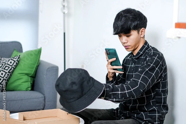 Fototapeta Serious Young Person Sits On The Floor Using His Smartphone To Capture Image Of New Bucket Hat