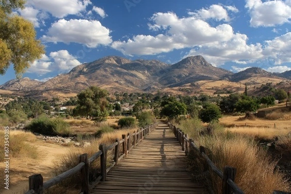 Fototapeta Wooden bridge leading to a valley nestled between mountains under a partly cloudy sky