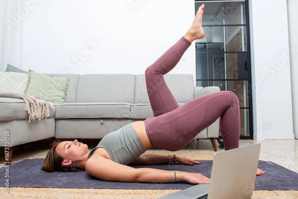 Fototapeta Young woman doing single leg bridge exercise at home with laptop guidance