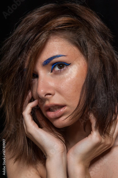 Fototapeta Unretouched portrait of a young pretty caucasian brunette girl posing in a studio on black background with soft light. She has messy hair and bright blue eye liner and beige lip gloss.