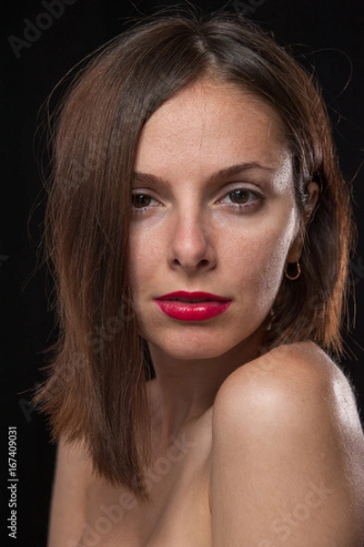 Fototapeta Unretouched, raw studio portrait of a young and pretty caucasian brunette girl with red lipstick, shot of black background with backlight.