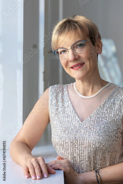 Obraz Portrait of smiling woman in festive clothes against a gray wall in a bright sunny room. Vertical photo