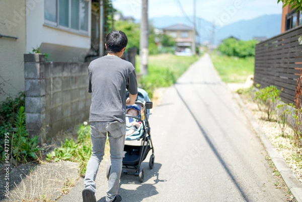Fototapeta A man is pushing a stroller with a baby in it. The baby is wearing a blue shirt. The man is walking down a street with a sidewalk