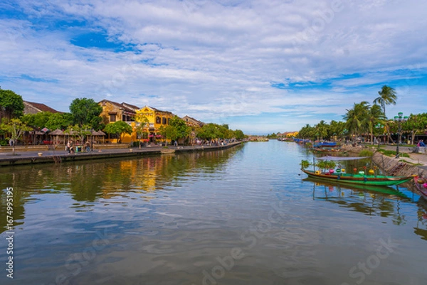 Fototapeta A busy riverbank scene in Hoi An, with numerous colorful tourist boats docked in the foreground and a row of classic yellow buildings in the background.