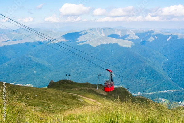 Fototapeta A view to red open cableway cabin over the top of the mountain and beautiful landscapes with blue mountains, green walley, Bucegi natural park, Sinaia, Romania.