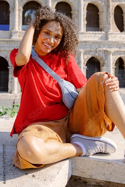 Fototapeta A joyful young woman with curly hair poses in a vibrant red shirt and tan shorts, set against ancient architecture, embodying travel and lifestyle themes of exploration and enjoyment