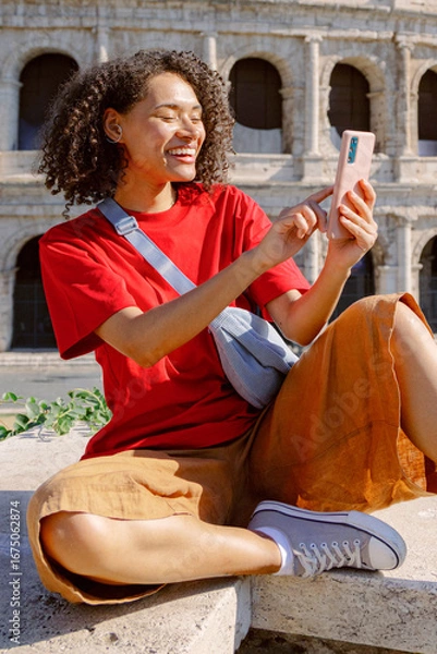 Fototapeta A cheerful young woman sits on a stone ledge, happily using her smartphone, enjoying the beautiful view of the ancient Colosseum in Rome, blending rich culture with modern technology