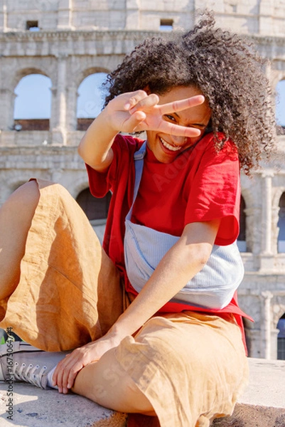 Fototapeta A cheerful and lively young woman poses at the iconic Colosseum, proudly showcasing her stylish outfit and making a playful peace sign gesture to capture the joyful ambiance of her experience