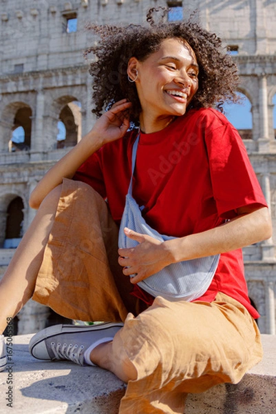 Fototapeta A stylish young person beams happily while wearing a bright red tshirt paired with comfy beige shorts, seated near the famous Colosseum, capturing a joyful and vibrant moment in time