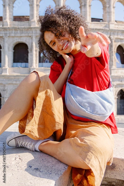 Fototapeta A joyful young woman poses playfully against the backdrop of the iconic Colosseum, proudly showcasing her stylish outfit and vibrant personality while capturing the moment in a delightful selfie