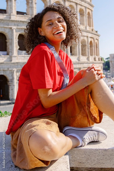 Fototapeta A joyful young woman in a vibrant red shirt and stylish brown skirt sits happily in front of the magnificent Colosseum, embracing the essence of travel and unforgettable adventure