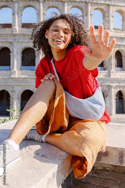 Fototapeta A joyful young woman waves at the camera while sitting on a stone ledge, with the Colosseum in the backdrop, showcasing her vibrant personality during her sightseeing adventure in Rome