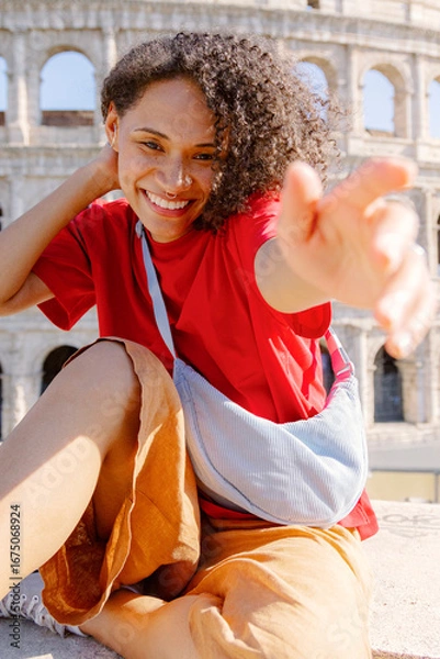 Fototapeta A cheerful young woman with curly hair beams a warm smile while casually sitting by the magnificent Colosseum, capturing a joyful moment full of adventure and excitement in the heart of Rome