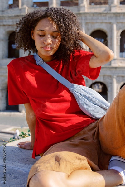 Fototapeta A stylish woman in a red tshirt and beige shorts relaxes near the iconic Colosseum in Rome, embodying a summer vibe that captures the essence of leisure in this culturally rich destination