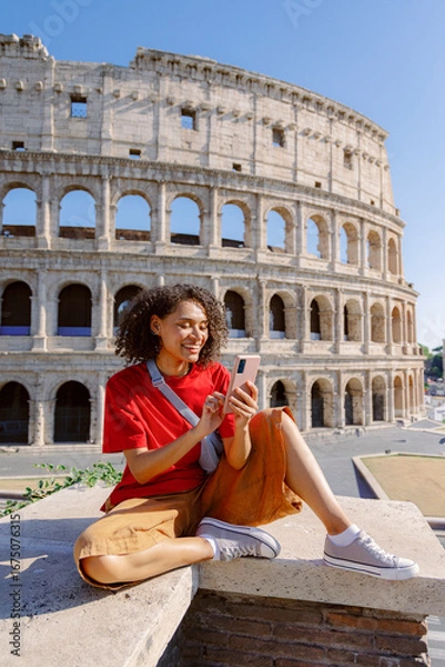 Fototapeta A young woman in stylish summer attire smiles as she relaxes on the sunlit steps of the Colosseum, focused on her smartphone while enjoying the lively atmosphere of Romes streets