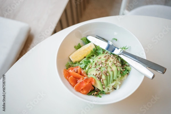 Fototapeta Close-up of  a bowl with green salad with smoked salmon, lettuce and avocado, garnished with seeds and lemon.