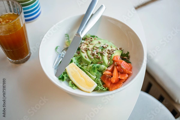 Fototapeta Overhead view of a bowl with green salad with smoked salmon, arugula and avocado, garnished with seeds and lemon, on a white table.