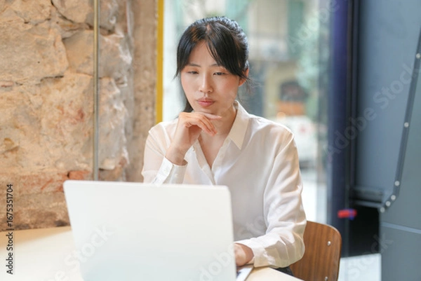 Fototapeta A woman is sitting at a table with a laptop in front of her. She is looking at the screen and she is deep in thought