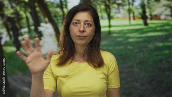 Obraz Middle aged hispanic woman wearing a yellow tshirt stands among green trees under sunlight and waves her hand in forest clearing; friendly greeting.