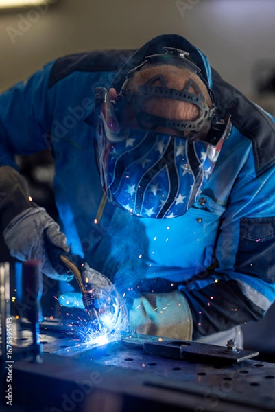 Fototapeta Man with welding mask in safety suit with gloves welds steel in home workshop. Welding flame with sparks. Dangerous work