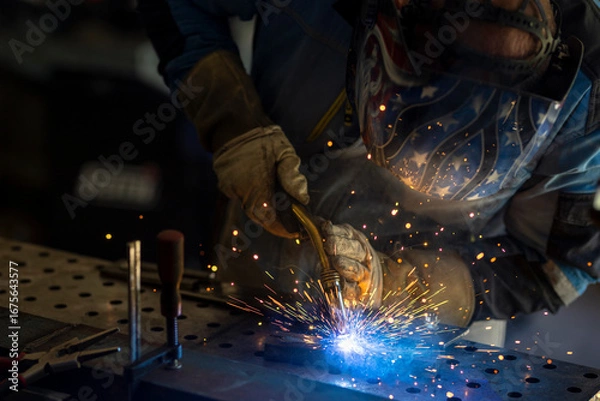 Fototapeta Man with welding mask in safety suit with gloves welds steel in home workshop. Welding flame with sparks. Dangerous work