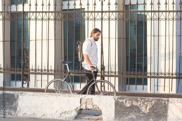 Obraz Young bearded ciclyst with black bicycle walking on the bridge in white shirt and leather backpack 