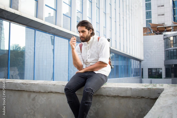 Fototapeta handsome guy with beard smocking cigarette on urban background with blue and gray glass  