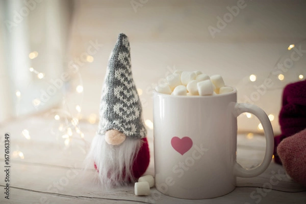 Fototapeta Christmas still life with white heart cup filled with chocolate and marshmallows, gnome doll and string lights. Festive, cozy  background with copy space.