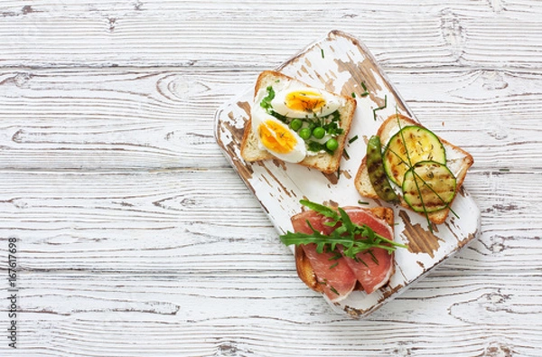 Fototapeta Toasts with eggs, ham and vegetables served on a white wood cutting board