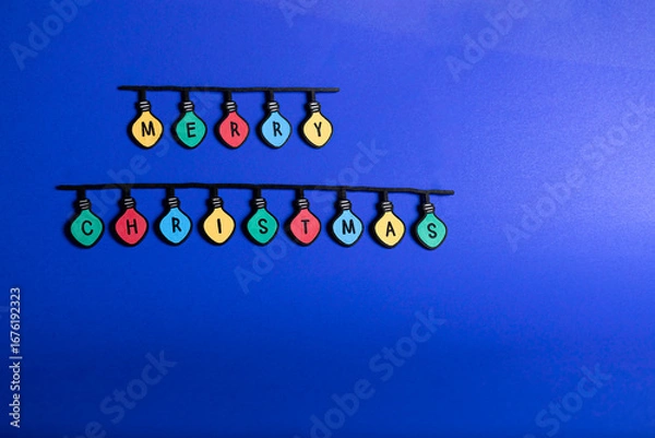 Fototapeta Christmas garland of colored light bulbs spelling out “Merry Christmas” on a blue background. Copy space